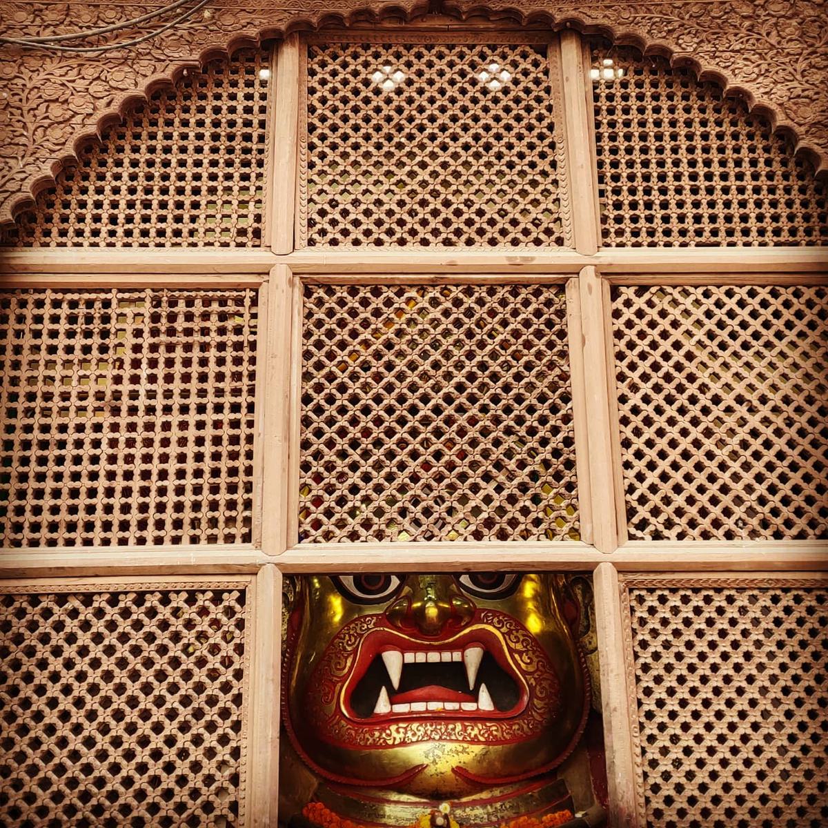 Bhairav's gilded face watching from behind a carved lattice window in Kathmandu